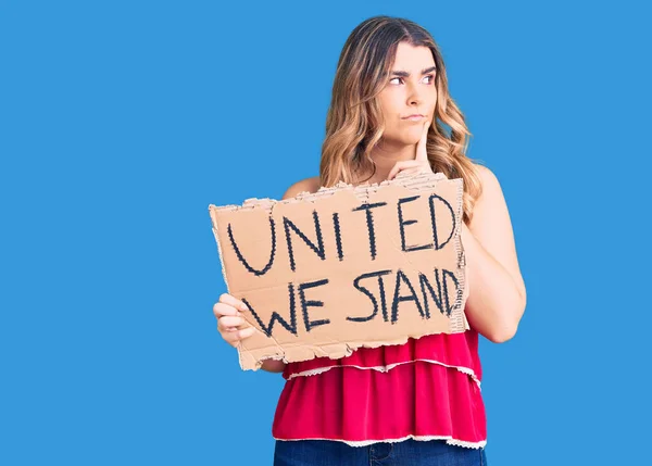 Young caucasian woman holding united we stand banner serious face thinking about question with hand on chin, thoughtful about confusing idea