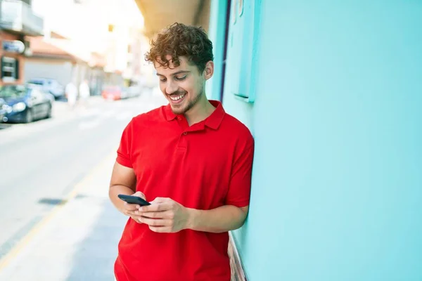 若いハンサムな白人男性は幸せな笑顔 市内のスマートフォンを使って壁にもたれる — ストック写真