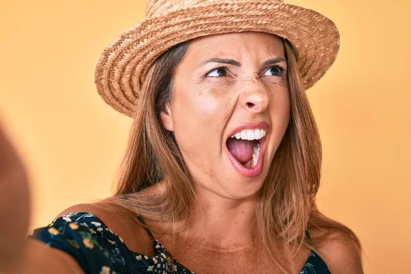 Middle age hispanic woman wearing summer hat taking a selfie angry and mad screaming frustrated and furious, shouting with anger. rage and aggressive concept.