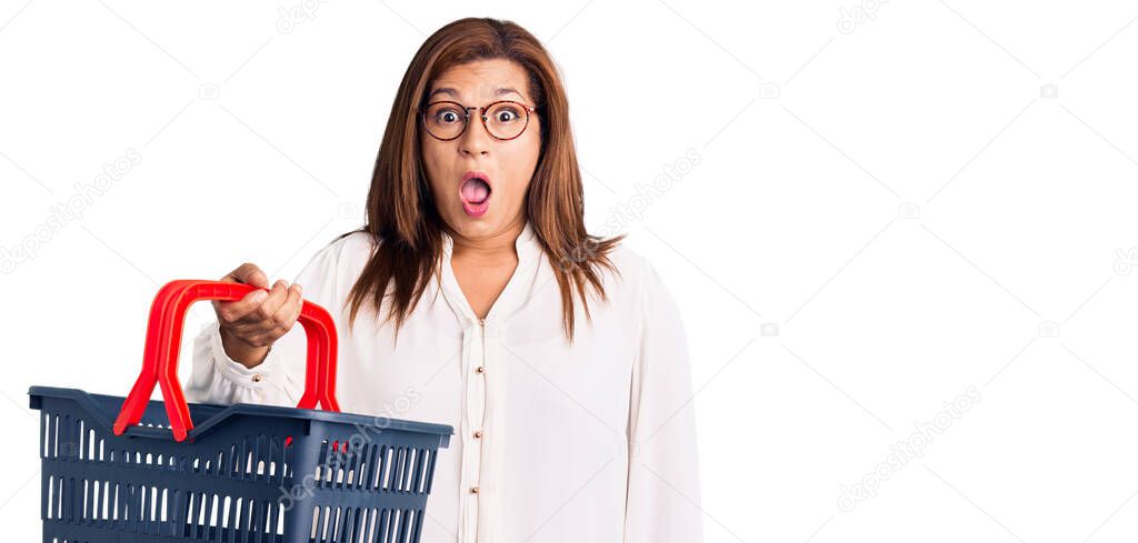 Middle age latin woman holding supermarket shopping basket scared and amazed with open mouth for surprise, disbelief face 