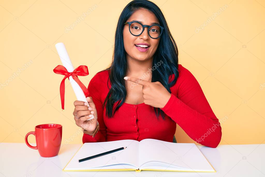 Beautiful latin young woman with long hair holding graduate degree diploma smiling happy pointing with hand and finger 