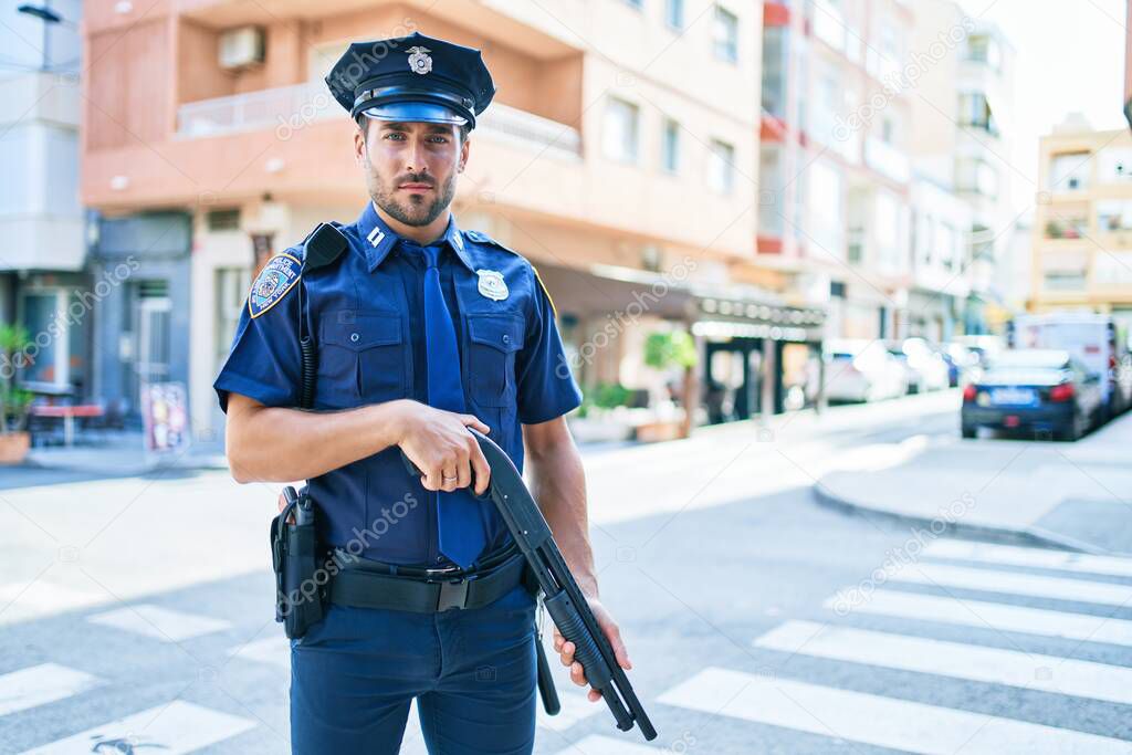 Young handsome hispanic policeman wearing police uniform with serious expression holding shotgun at town street.