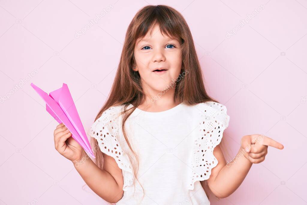 Little caucasian kid girl with long hair holding paper airplane smiling happy pointing with hand and finger to the side 