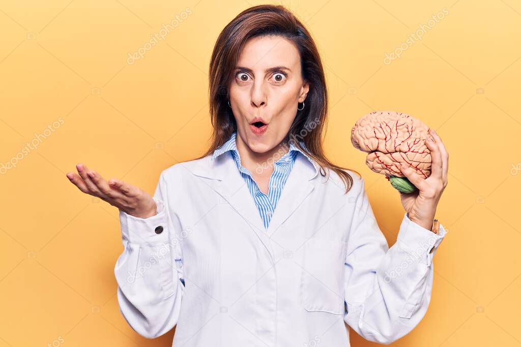 Young beautiful woman wearing doctor coat holding brain scared and amazed with open mouth for surprise, disbelief face 