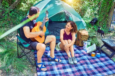 Ormanda kamp yaparken gülümseyen orta yaşlı bir çift. Yerde oturup klasik gitar çalarak şarkı söylemek.