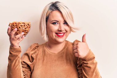 Güzel ve iri bir kadın elinde Alman krakerleriyle beyaz arka planda gülümseyen, mutlu ve pozitif gülümseyen, başparmağını kaldıran mükemmel ve onay işareti yapan.