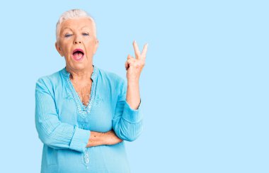 Mavi gözlü, gri saçlı, yaz elbisesi giyen, kameraya gülümseyen ve zafer işareti yapan son sınıf güzeli bir kadın. İki numara.. 