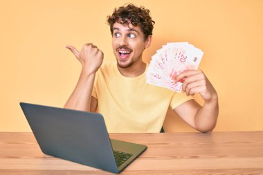 Masaya kıvırcık saçlı genç beyaz bir adam oturmuş dizüstü bilgisayarla çalışıyor ve elinde İsrail şekeri banknotları ile baş parmağını kaldırıp ağzı açık bir şekilde gülümsüyor. 