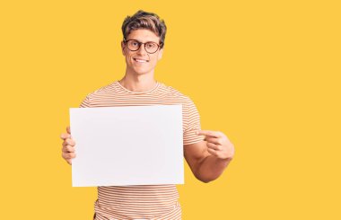 Boş afiş tutan genç yakışıklı adam mutlu bir şekilde el ve parmağıyla işaret ediyor. 