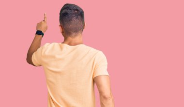 Genç, yakışıklı, sıradan kıyafetler giyen, geriye doğru poz veren, parmaklarıyla öne bakan bir adam. 