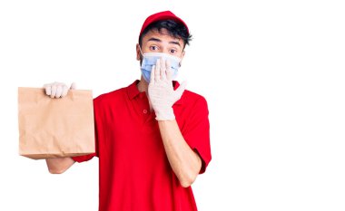 Teslimat üniforması giyen ve tıbbi maske takan, ağzı elle kapatılmış, şok olmuş ve hata yapmaktan korkan genç İspanyol bir adam. şaşırmış ifade 