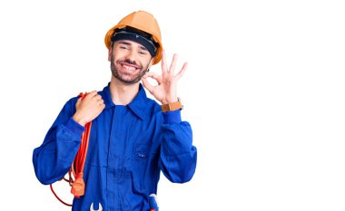 Genç İspanyol adam, elektrokrat üniforması giyiyor. Kablo tutuyor. Parmaklarıyla imza atıyor, gülümsüyor. 