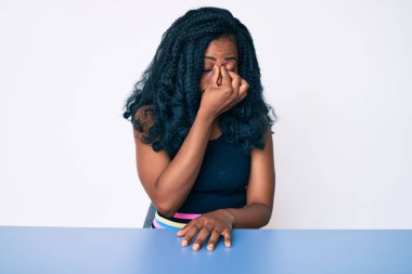 Güzel Afrikalı kadın gündelik giysiler giyip masada oturuyor. Yorgun, burnu ve gözleri ovuşturuyor. Yorgunluk ve baş ağrısı çekiyor. Stres ve hüsran kavramı. 