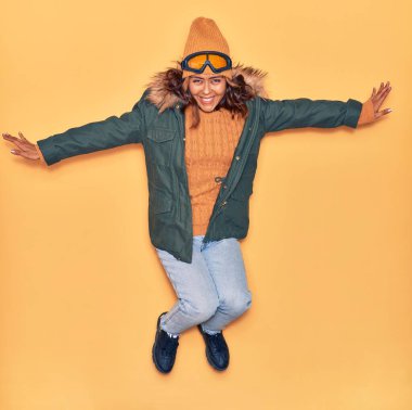 Kışlık giysiler giyen genç, güzel Latin kadın mutlu gülümsüyor. Yüzünde ve kollarında tebessümle, izole edilmiş sarı arka planda atlıyor.