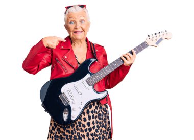 Son sınıf güzeli, mavi gözlü, gri saçlı modern görünümlü, elektro gitar çalan neşeli gülümseyen ve parmaklarını dişleriyle ve ağzıyla işaret eden bir kadın. diş sağlığı kavramı. 