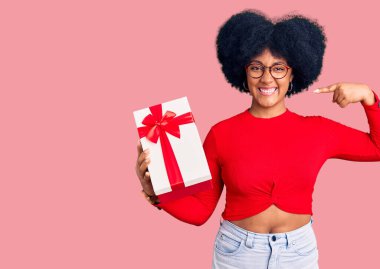 Genç Afrikalı Amerikalı kız elinde hediyeyle neşeli bir şekilde gülümseyip parmaklarını ve ağzını işaret ediyor. diş sağlığı kavramı. 