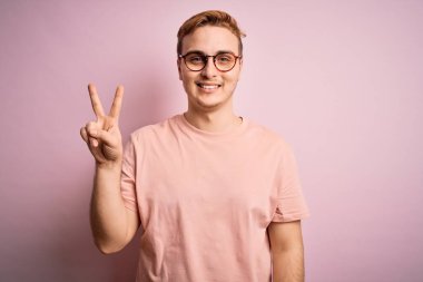 Genç, kızıl saçlı, günlük tişört giyen, izole edilmiş pembe arka planda duran, parmaklarını zafer işareti yaparken gösteren kameraya gülümseyen bir adam. İki numara..
