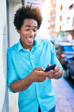 Genç, yakışıklı, Afro-Amerikan bir adam günlük giysiler giyip mutlu gülümsüyor. Yüzünde gülümsemeyle duvara yaslanıyorsun. Şehir caddesinde akıllı telefon kullanıyorsun..