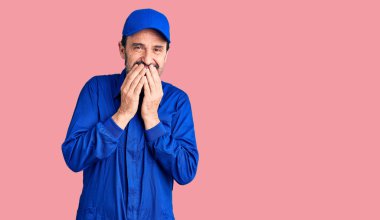 Tamirci üniforması giyen orta yaşlı yakışıklı adam gülüyor ve utanıyor kıkırdıyor elleriyle ağzını kapatıyor, dedikodu ve skandal konsepti yapıyor. 