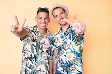 Yaz şapkası ve Hawaii tişörtü giymiş iki genç eşcinsel, iki elini de zafer işareti yaparken göstererek gülümsüyor. İki numara.. 