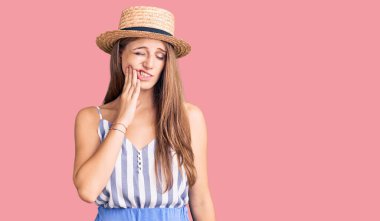 Sarışın, yaz şapkası takan genç bir kadın diş ağrısı ya da diş hastalığından dolayı acı dolu bir ifadeyle ağıza dokunuyor. dişçi 