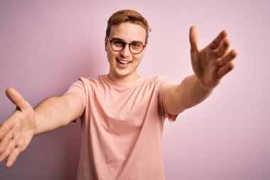 Genç, kızıl saçlı, günlük tişört giyen, izole edilmiş pembe arka plan üzerinde duran ve kucaklaşmak için kollarını açmış gülümseyen kameraya bakan yakışıklı bir adam. Mutluluğu kucaklayan neşeli bir ifade..