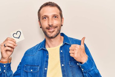 Elinde kalp taşıyan yakışıklı adam gülümseyen mutlu ve pozitif, başparmak yukarı mükemmel ve onay işareti yapıyor. 