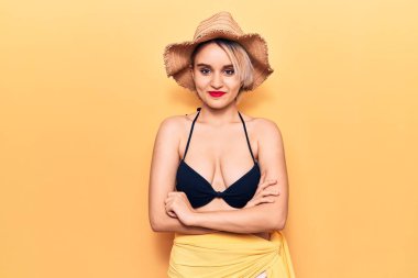 Bikini giyen güzel sarışın bir kadın ve kollarını kavuşturup kameraya bakan mutlu yüzlü bir yaz şapkası. pozitif kişi. 