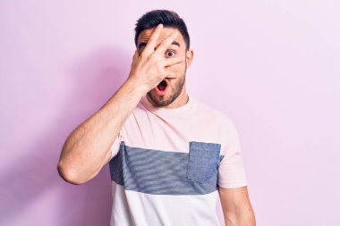 Sakallı, sıradan tişörtlü, pembe arka planda duran, yüzü ve gözleri kapalı, parmaklarından korkan genç yakışıklı bir adam.