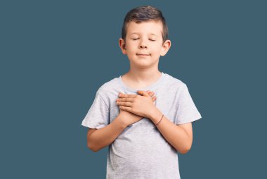 Hoş sarışın çocuk, sıradan kıyafetler giyiyor, elleri göğsünde, gözleri kapalı gülümsüyor ve yüzünde minnet ifadesi var. sağlık kavramı. 