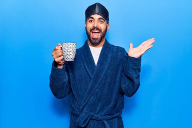 Uyku maskesi takan ve sabahlık giyen genç İspanyol adam zaferi kutlamak için kahveyi yudumlarken mutlu bir gülümseme ve elini kaldırarak kazanan yüz ifadesi takınıyor. 