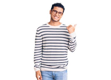 Gündelik kıyafetler ve gözlüklerle gülümseyen, gülümseyen ve başparmağıyla yüzünü gösteren İspanyol asıllı yakışıklı genç adam.. 