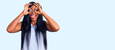 Genç Afro-Amerikalı kadın günlük kıyafetler giyiyor dürbün gibi jest yapıyor, dili dışarı çıkarıyor, gözleri parmaklarından bakıyor. çılgın ifade. 