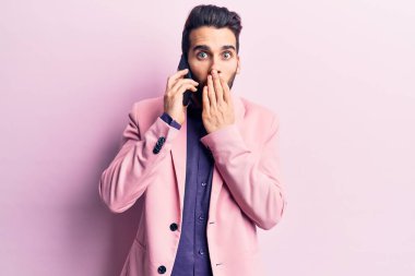 Sakallı genç yakışıklı adam akıllı telefonda konuşuyor ağzını eliyle kapatıyor, şaşırmış ve hata yapmaktan korkuyor. şaşırmış ifade 