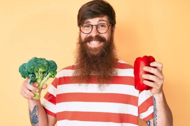 Uzun sakallı, elinde brokoli ve yüzünde mutlu ve havalı bir gülümsemeyle gülümseyen kırmızı biberli yakışıklı genç bir adam. diş gösterme. 