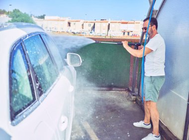 Orta yaşlı yakışıklı, günlük kıyafetler giyen ve mutlu gülümseyen güneş gözlüklü bir adam. Yüzünde gülümsemeyle ayakta durup su tabancası kullanarak araba yıkamak..