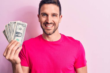 Soyutlanmış pembe arka planda bir deste para tutan yakışıklı genç adam pozitif ve mutlu görünüyor ve kendine güvenen bir gülümsemeyle dişlerini gösteriyor.