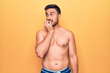 Sarı arka planda kolsuz tişört giyen sakallı yakışıklı genç adam gergin görünüyor ve elleri ağızdan tırnak yiyor. Endişe sorunu.