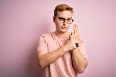 Genç, yakışıklı, günlük tişört giyen, pembe arka planda duran, el hareketleriyle sembolik silah tutan, cinayet silahlarıyla oynayan, öfkeli yüzlü bir adam.