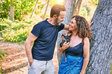 Orta yaşlı çift gülümsüyor. Chihuahua köpeği kucağında. Ormandaki kameraya bakıyor.