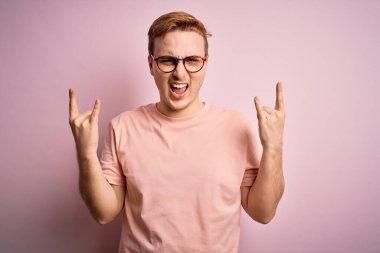 Genç, kızıl saçlı, günlük tişört giyen, izole edilmiş pembe arka plan üzerinde duran çılgın bir ifadeyle elleri havada rock sembolü yapan genç bir adam. Müzik yıldızı. Ağır müzik konsepti.