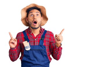 Yakışıklı Latin Amerikalı, tamirci üniforması giyen genç adam şaşkın ve şaşırmış bir şekilde yukarı bakıyor ve parmaklarıyla işaret ediyor ve kollarını kaldırıyor.. 