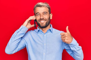 Sakallı yakışıklı sarışın genç adam akıllı telefonla konuşuyor mutlu ve olumlu gülümsüyor, başparmağıyla mükemmel ve onay işareti yapıyor.