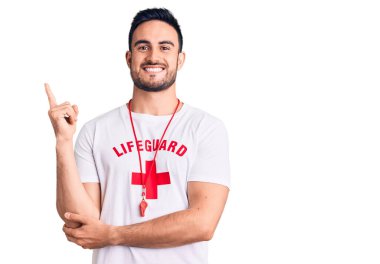 Yüzünde kocaman bir gülümseme olan cankurtaran üniforması giyen genç yakışıklı bir adam, kameraya el ve parmak ile bakıyor.. 