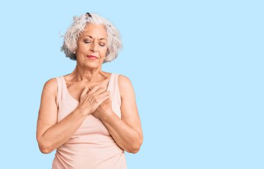 Üstün gri saçlı, günlük kıyafetler giyen, elleri göğsünde, gözleri kapalı gülümseyen ve yüzünde minnet ifadesi olan bir kadın. sağlık kavramı. 