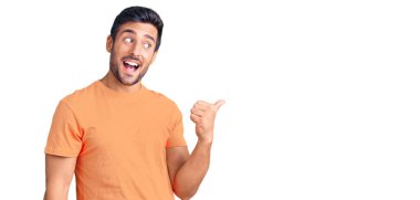 Gündelik kıyafetler giyen, gülümseyen, gülümseyen ve başparmağıyla yan tarafı gösteren İspanyol genç adam.. 