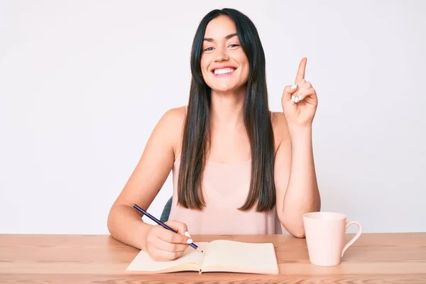 Young caucasian woman sitting at the desk writing book drinking coffee surprised with an idea or question pointing finger with happy face, number one