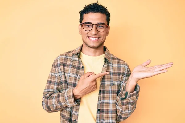手で提示しながらカジュアルな服や眼鏡を身に着けている若いハンサムなヒスパニック系の男は驚きとカメラに笑みを浮かべて指で指して — ストック写真