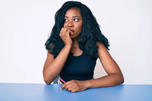 漂亮的非洲女人穿着休闲装坐在桌子上 看上去很紧张 双手放在咬指甲的嘴上 焦虑问题 — 图库照片