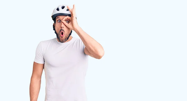 Young handsome man wearing bike helmet doing ok gesture shocked with surprised face, eye looking through fingers. unbelieving expression.